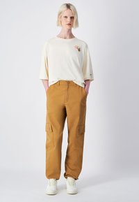 Camiseta oversized crema con un pequeño gráfico en el pecho, combinada con pantalones cargo marrones con bolsillos laterales, llevados con zapatillas blancas.