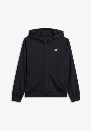 Garçon debout portant un sweat à capuche noir zippé, une chemise grise à motifs, un pantalon noir et des baskets noir et blanc sur un fond gris uni.