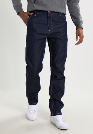 Mand iført mørkeblå denimjeans med hvide syninger, grå sweatshirt og hvide sneakers, står med den ene hånd i lommen.