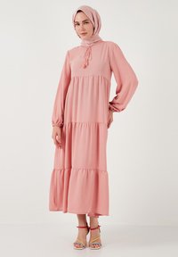 Robe longue rose à volants en tissu léger, avec des manches longues et un col à cordon, assortie de sandales colorées à brides.