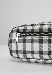 Sac à carreaux noir et blanc avec une surface texturée, doté d'une fermeture éclair argentée et d'une forme rectangulaire. La surface a une finition brillante.