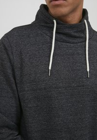 Hanorac gri închis cu gâtul înalt. Fabricat dintr-un material texturat, are șnururi albe ajustabile și capete din metal argintiu. Design casual.