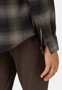 Camisa de franela a cuadros en tonos de gris y negro con puños abotonados. Mostrada junto a pantalones de textura marrón oscuro.