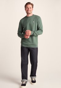 Smiling man stands wearing green sweatshirt, dark rolled-up jeans, and black sneakers with white soles against a plain beige background.