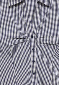 Chemise à carreaux bleu et blanc avec boutons, détail froncé au niveau de la poitrine et col pointu.