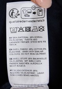 Pesulaputket valkoisella kankaalla, jossa yksityiskohtia materiaaleista: 95% puuvillaa, 5% elastaania. Sisältää pesusymboleita ja monikielisiä ohjeita.