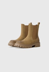 Botas de tobillo de ante marrón con paneles laterales elásticos, punta redonda y suela de goma gruesa. Cuenta con una lengüeta para ponerse fácilmente.