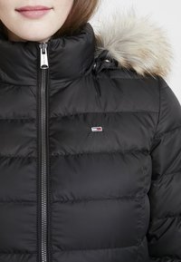 Black puffy jacket with a high collar and faux fur trim on the hood. Features a silver zip and small embroidered logo on the chest.