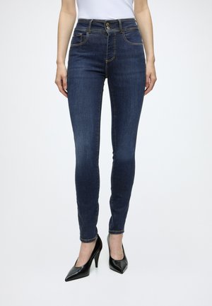 Kvinde iført mørkeblå skinny jeans med dobbelte knapper i taljen og sorte højhælede sko, stående mod en ensfarvet baggrund.