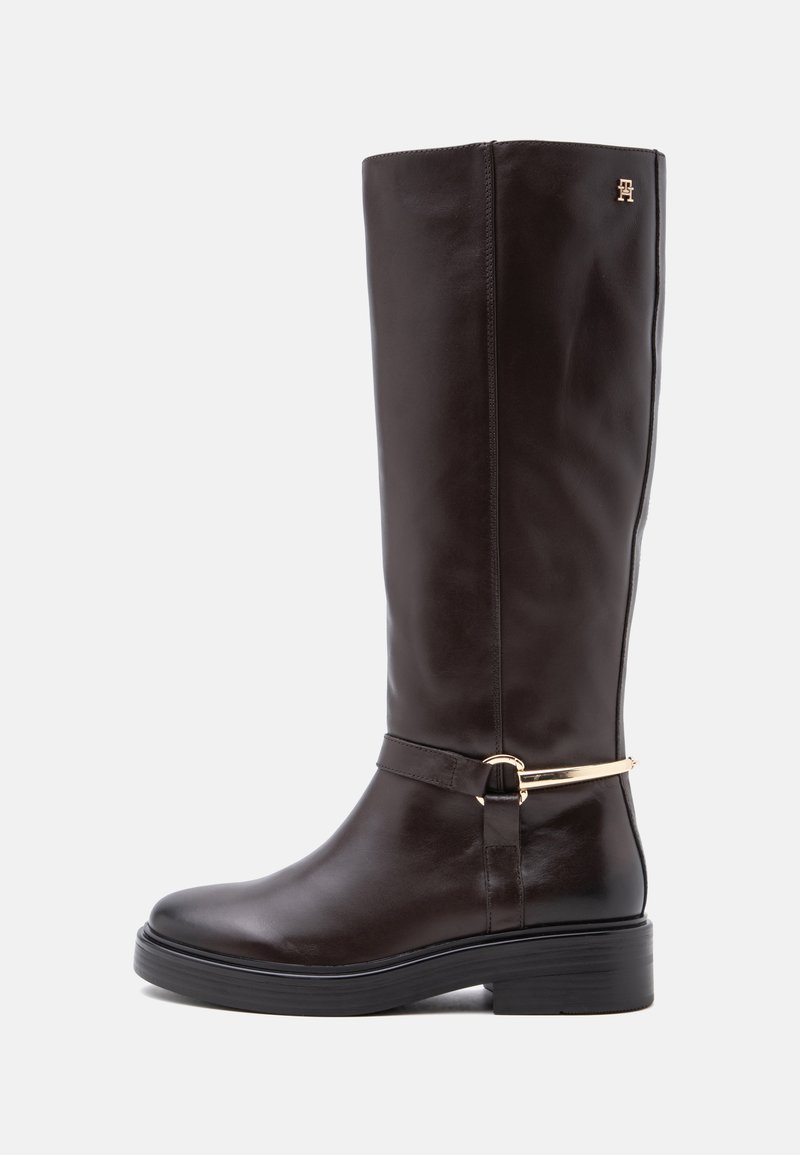 Botte haute en cuir marron foncé avec un petit talon, une sangle décorative dorée et un emblème près du haut.