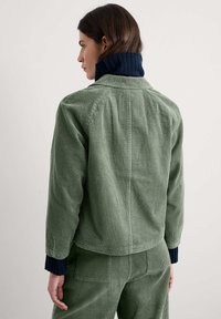 Vrouw in een groene corduroy jas en broek met een marineblauwe geribbelde coltrui, gezien van achteren tegen een effen achtergrond.