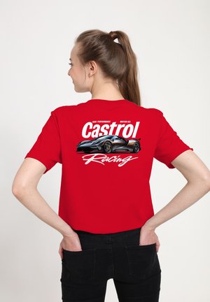Młoda kobieta z brązowymi włosami związanymi w kucyk, ubrana w czerwony t-shirt z czarnym samochodem sportowym i napisem "Castrol Racing" na plecach.