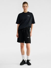Junge Frau, die ein schwarzes Oversized-T-Shirt und Shorts mit weißen hohen Socken und schwarzen Sneakers auf hellem einfarbigem Hintergrund trägt.