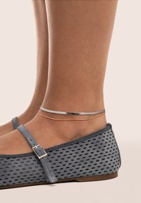 Zapatos planos de ante gris presentan un diseño perforado y una correa con hebilla. Se pueden ver dos finas pulseras metálicas en los tobillos.