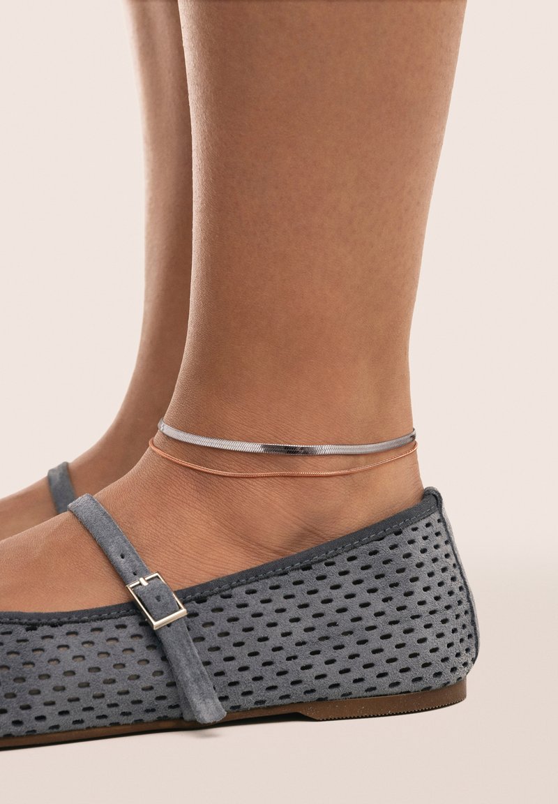 Zapatos planos de ante gris presentan un diseño perforado y una correa con hebilla. Se pueden ver dos finas pulseras metálicas en los tobillos.