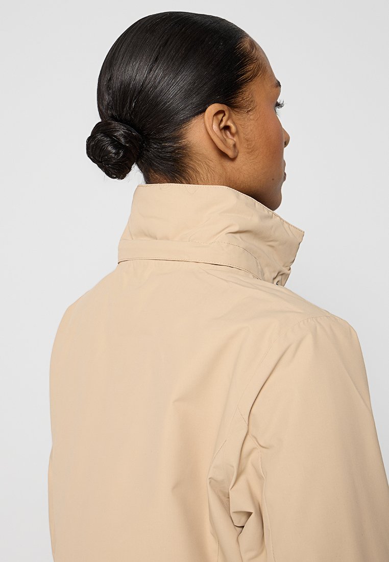 Frau mit dunklem Haar in einem tiefen Dutt, trägt eine beige Jacke mit hohem Kragen, von hinten und zur Seite gegen einen schlichten Hintergrund betrachtet.