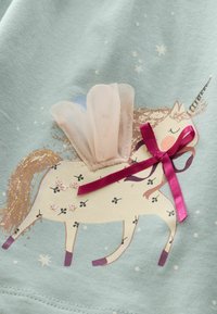 Tissu bleu clair avec un graphique de licorne. La licorne a une crinière dorée, un nœud rose, un accent en tulle et des fleurs décoratives. Des étoiles scintillantes sont visibles.