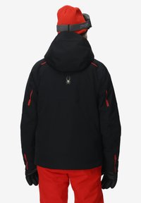 Schwarze Skijacke mit roten Akzenten, Kapuze und Spinnenlogo auf dem Rücken. Getragen mit roten Hosen und schwarzen Handschuhen. Mattes Finish, isoliertes Design.
