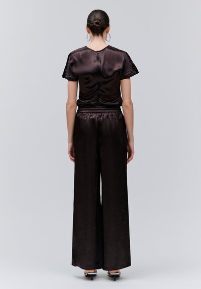 Femme aux cheveux relevés en chignon, portant un haut noir en satin à manches courtes et un pantalon large, debout de dos sur un fond gris clair.