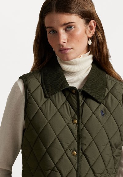 Gestepptes olivgrünes Westchen mit einem Kordkragen, Messingdruckknöpfen und gesticktem Logo. Über einem weißen gerippten Rollkragenpullover getragen.