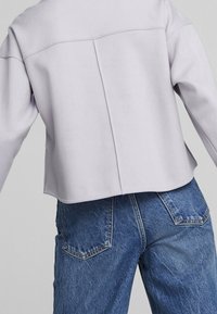 Giacca cropped di un leggero colore viola con taglio dritto e maniche leggermente oversize, abbinata a jeans in denim blu a gamba tapered con tasche posteriori.