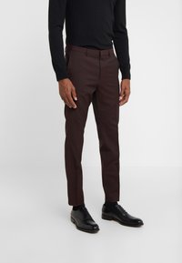 Pantaloni marroni slim fit realizzati in un tessuto liscio, con frontale piatto, tasche laterali e aperture per le gambe strette. Abbinati a scarpe eleganti nere.