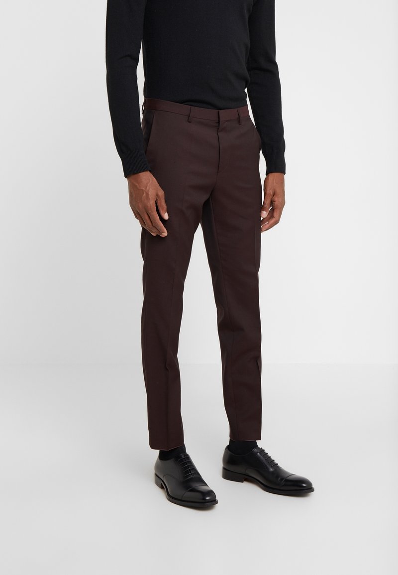 Pantaloni marroni slim fit realizzati in un tessuto liscio, con frontale piatto, tasche laterali e aperture per le gambe strette. Abbinati a scarpe eleganti nere.