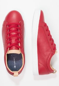 Par av röda lädersneakers med vita sulor, röda snören, beige detaljer och synlig Lacoste-logotyp på blårandiga innersulor.