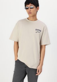 Camiseta oversize beige, de manga corta, con el texto gráfico en navy "ALABAMA Rodeo." Combinada con pantalones gris oscuro, el modelo muestra un atuendo casual.
