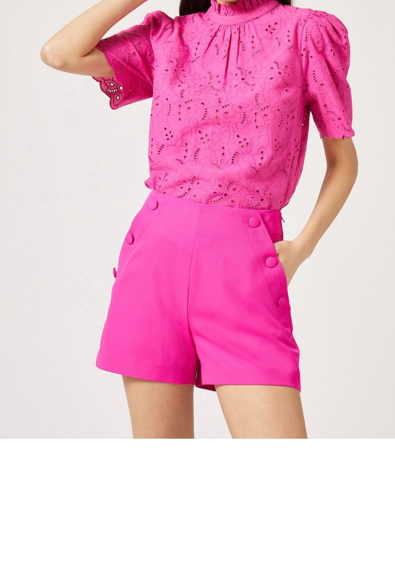 Blusa rosa brillante con design a occhiello e maniche a sbuffo, abbinata a pantaloni corti rosa a vita alta con grandi bottoni sui lati.
