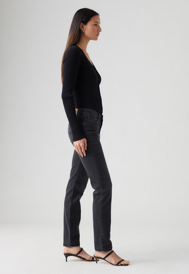 Cardigan long à manches longues noir côtelé avec des boutons, associé à un jean droit gris foncé et des sandales à talons noires, présenté de côté.