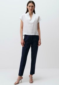 Weiße Leinenbluse mit V-Ausschnitt und kurzen Ärmeln, kombiniert mit dunkelblauen schmalen Hosen. Das Model trägt silberne Absatzsandalen.