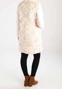 Faux-Fur-Mantel in Creme mit einer strukturierten, flauschigen Oberfläche. Lange Ärmel aus glattem Stoff. Schwarze Skinny-Jeans und braune Ankle-Boots runden den Look ab.