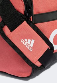 Borsa a tracolla corallo con una tracolla nera regolabile, dotata di chiusura a zip e di un prominente logo Adidas bianco. Tessuto testurizzato con accenti lisci.