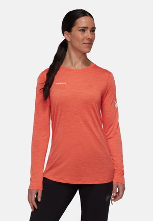 Kvinde med mørkt hår i en hestehale, iført en lyseorange langærmet skjorte og sorte bukser, stående foran en ensfarvet lys baggrund.