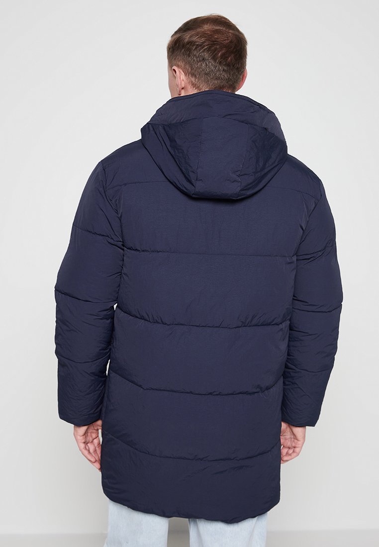 Veste rembourrée marine avec capuche, présentant un design matelassé, des manches longues et une coupe mi-longue. Texture lisse et construction simple.