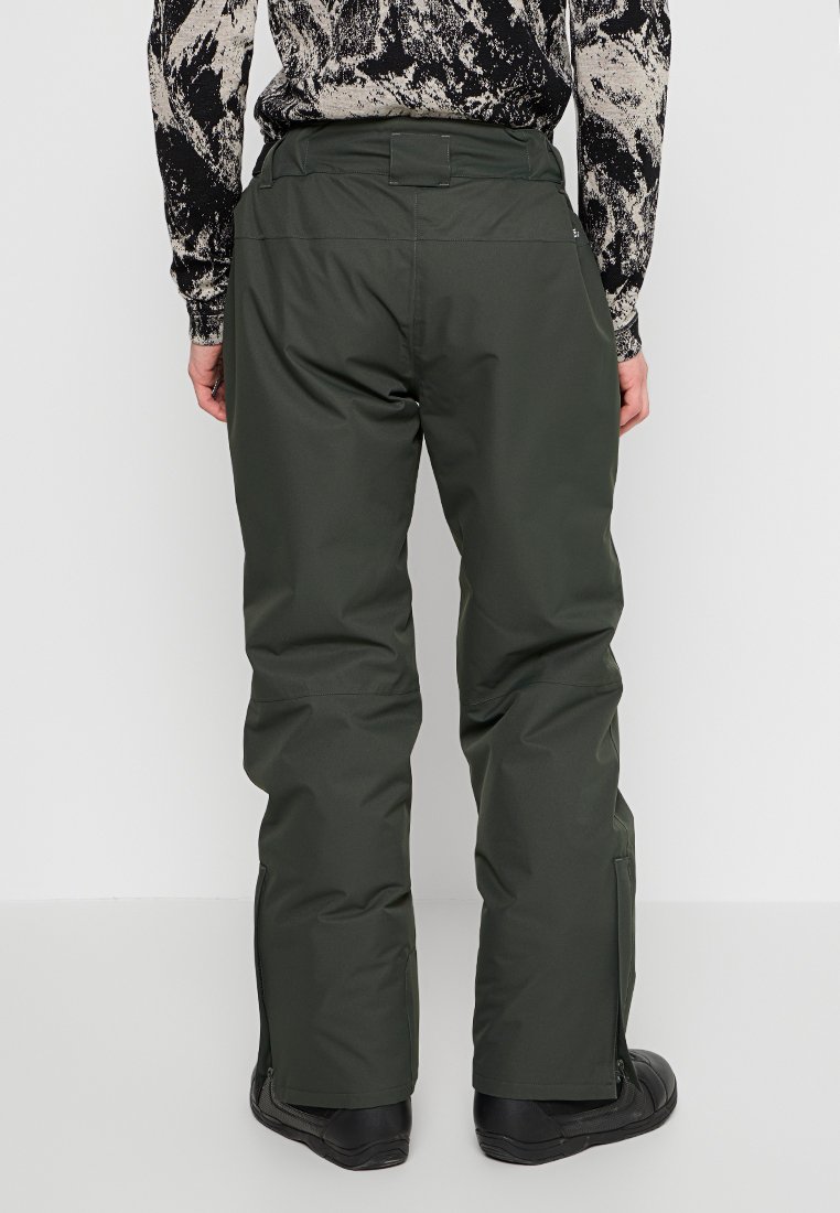 Eine Person trägt eine dunkelgrüne wasserdichte Hose und schwarze Stiefel, von hinten gegen einen schlichten weißen Hintergrund gezeigt.