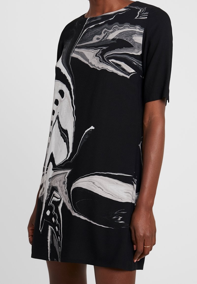 Robe noire à manches courtes avec motif abstrait marbré blanc et gris, portée par une personne à la peau foncée et portant une fine bague en or au doigt.