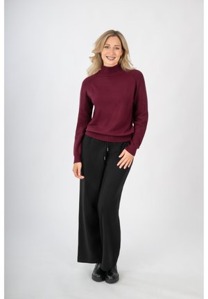 Burgundowa dzianinowa sweter z golfem połączony z luźnymi czarnymi spodniami. Modelka stoi na neutralnym tle, uśmiechając się.