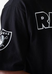 Juodas medvilnės marškinėlis su užrašu "RAIDERS" ryškiu baltu šriftu, su juodu ir baltu logotipu ant kairiojo rankovės. Minkšta, kvėpuojanti textura.