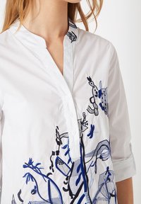 Blusa blanca con cuello mao, adornada con diseños florales y abstractos bordados en azul, botones en la parte delantera y mangas remangadas.
