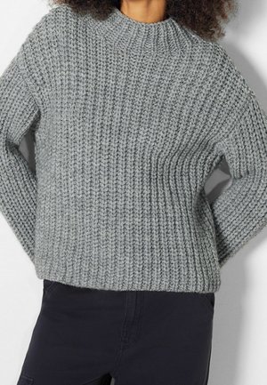 Personne portant un pull côtelé gris épais et un pantalon noir, debout les mains derrière le dos devant un fond uni.