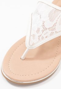 Valkoinen pitsiset flip-flop sandaali, jossa on thong-hihna, beige pehmustettu pohja ja hillitty metallinen yksityiskohta pohjan reunan ympärillä.