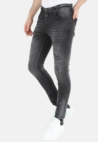 Grijze skinny jeans met verweerde accenten, waaronder scheuren en verfsplinters, gecombineerd met witte sneakers en een strakke witte shirt.
