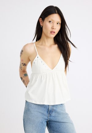 Jeune femme aux longs cheveux foncés portant un haut blanc sans manches avec des détails cloutés et un jean bleu clair, debout devant un fond uni.