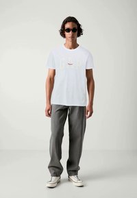Hvid bomulds T-shirt med "GAP Original" logo i beige og rød. Parret med grå, løstsiddende bukser og hvide sneakers.