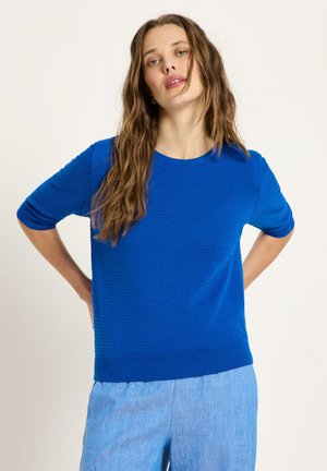 Woman with long brown hair wearing a bright blue ribbed short-sleeve sweater and light blue pants, standing with hands on hips.