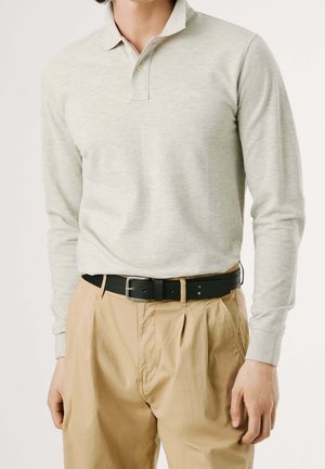 Mężczyzna ubrany w jasnoszarą koszulkę polo z długim rękawem, włożoną w beżowe plisowane spodnie z czarnym skórzanym paskiem, stojący na jasnym tle.