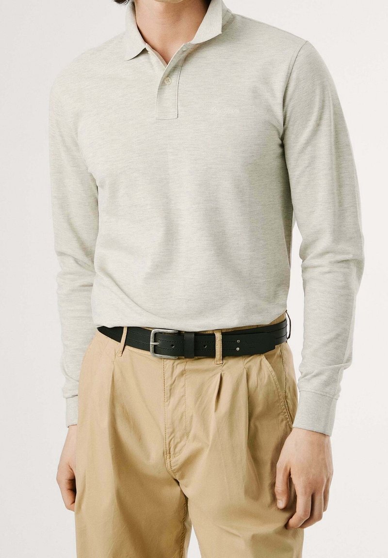 Homme portant un polo gris clair à manches longues, rentrée dans un pantalon plissé beige avec une ceinture en cuir noire, debout devant un fond clair.