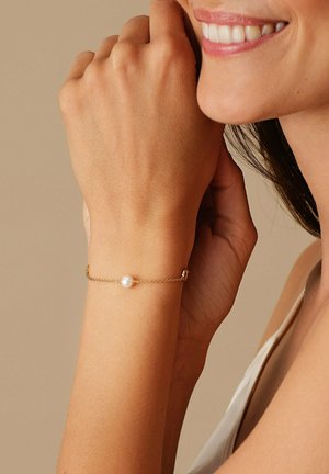 Bracciale in oro con un unico centro di perla bianca, indossato su una mano su uno sfondo neutro. Texture liscia con un design delicato.